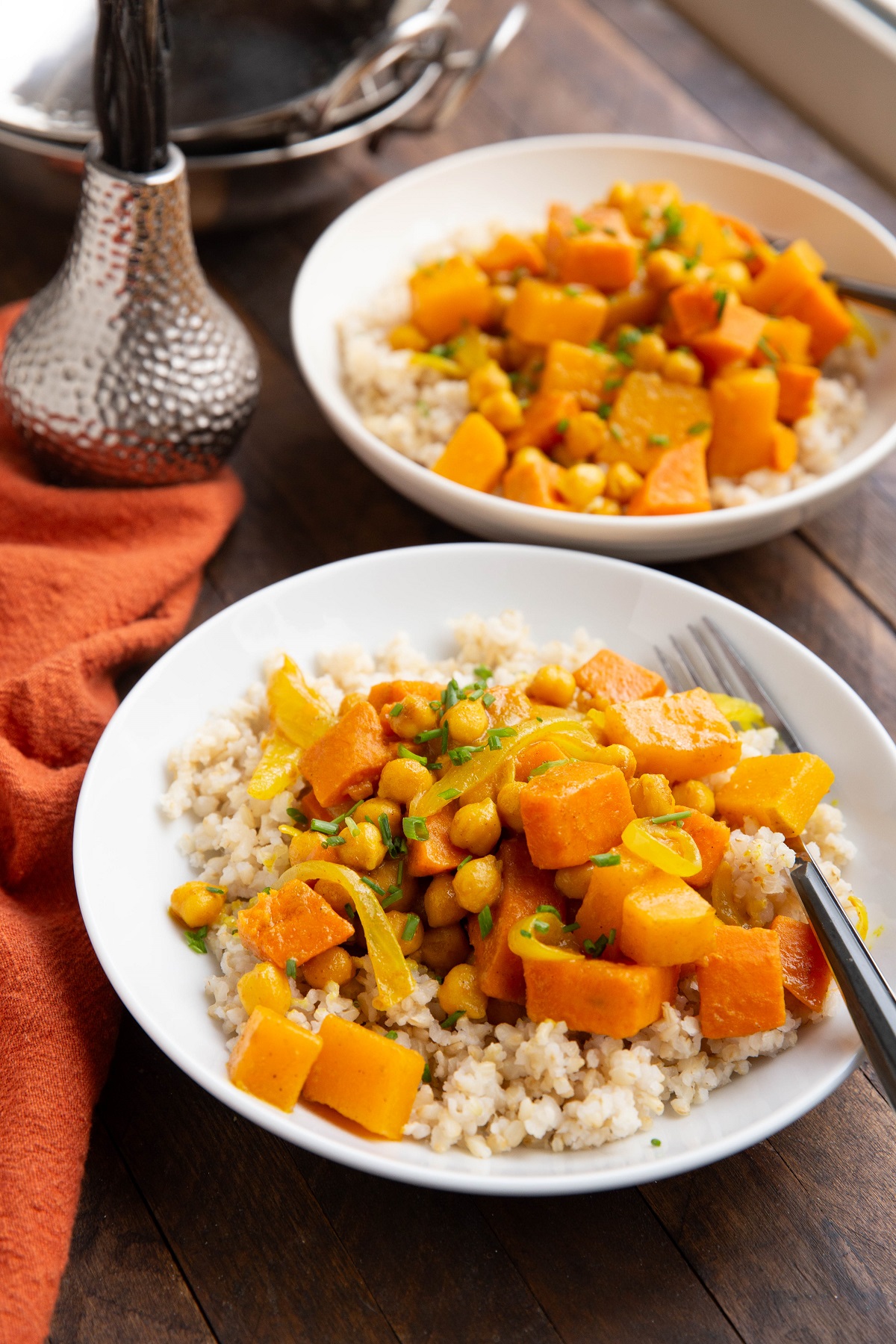 Butternut squash and yam curry recipe with brown rice and garbanzo beans in two white bowls on a wood background with a red napkin to the side.