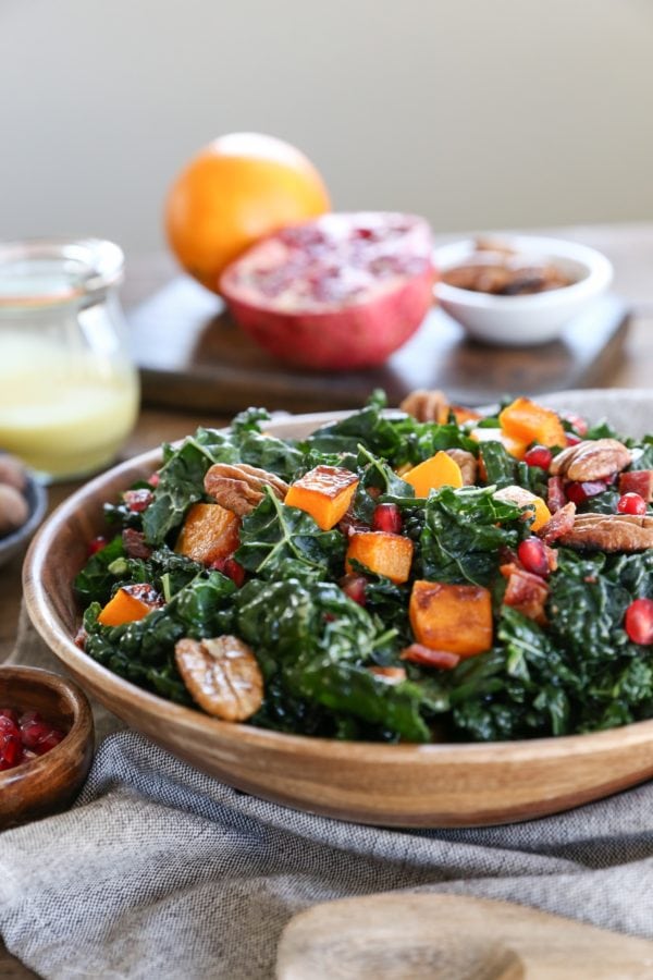 Wooden bowl of kale salad with roasted butternut squash, pecans, pomegranate seeds, and fresh ingredients in the background.