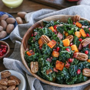 Butternut squash kale salad in a large wooden bowl with pecans and pomegranate seeds. A napkin and fresh ingredients all around.