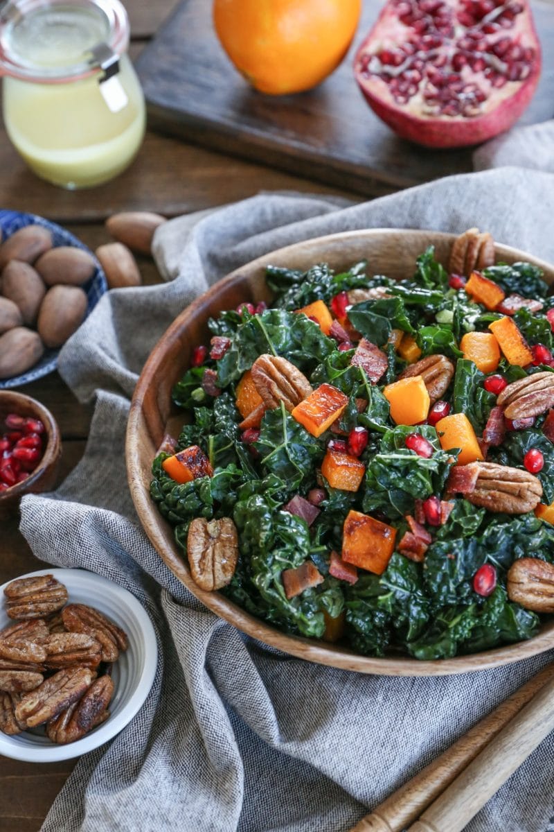 Butternut squash kale salad in a large wooden bowl with pecans and pomegranate seeds. A napkin and fresh ingredients all around.