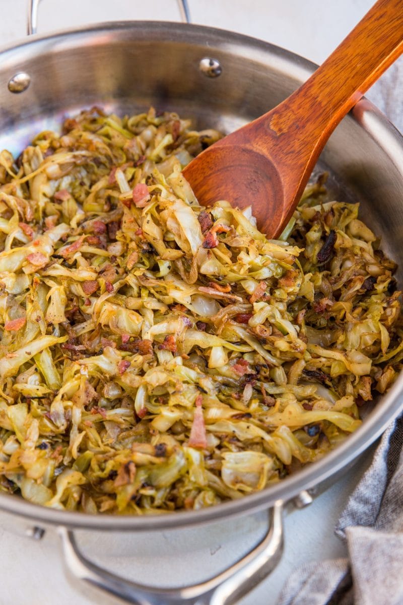 Stainless steel skillet full of caramelized cabbage with onions and bacon. A wooden spoon is ready to serve.