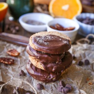 Four cardamom orange cookies stacked on top of each other with nuts and chocolate in the background.