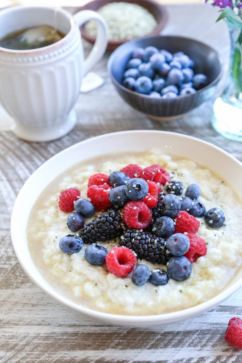 Cauliflower Porridge in a white bowl with milk, honey, fresh berries, and hemp hearts. a mug of tea and a bowl of blueberries in the background.