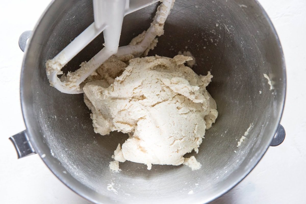 A metal mixing bowl containing a ball of cookie dough and a white paddle attachment from a stand mixer, perfect for preparing gluten-free cheese flatbread or trying your favorite pupusa recipe.