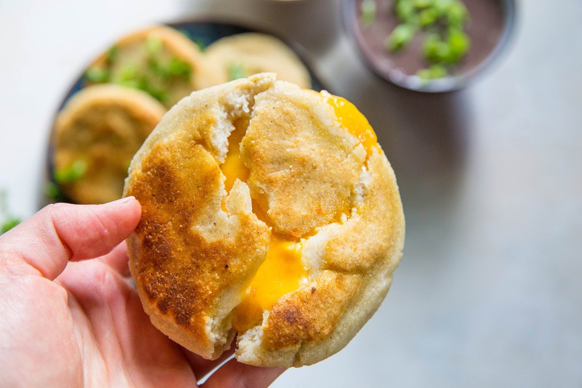 A blue plate with four golden, pan-fried cheese pupusas garnished with chopped green onions and cilantro. Salsa and refried beans are in bowls nearby, along with scattered herbs on a white surface—a tempting cheese pupusa recipe.