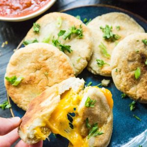 Hand pulling a cheese pupusa apart so you can see the gooey cheesy inside. A plate with more pupusas and a bowl of salsa and beans.