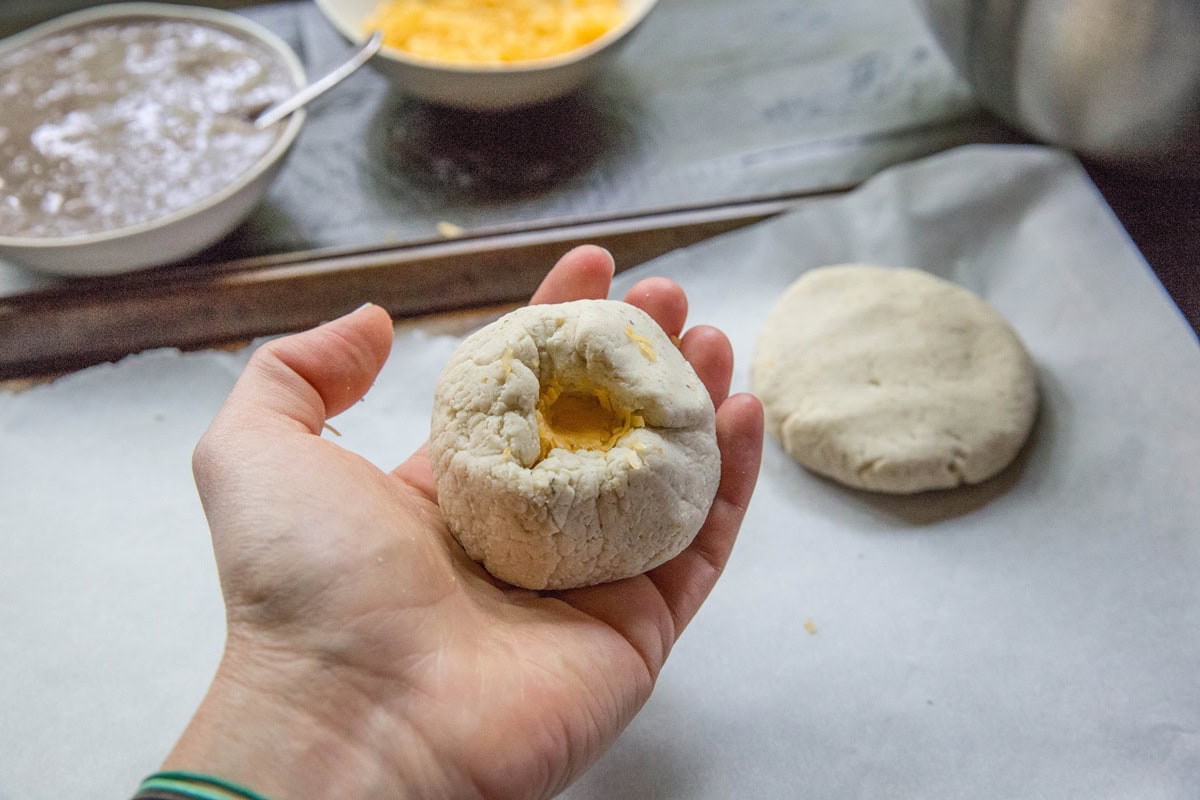 A hand holding an uncooked biscuit dough ball with a thumb-pressed indentation in the center, perfect for making cheese pupusas; another dough ball and bowls of ingredients are in the background on a baking sheet.