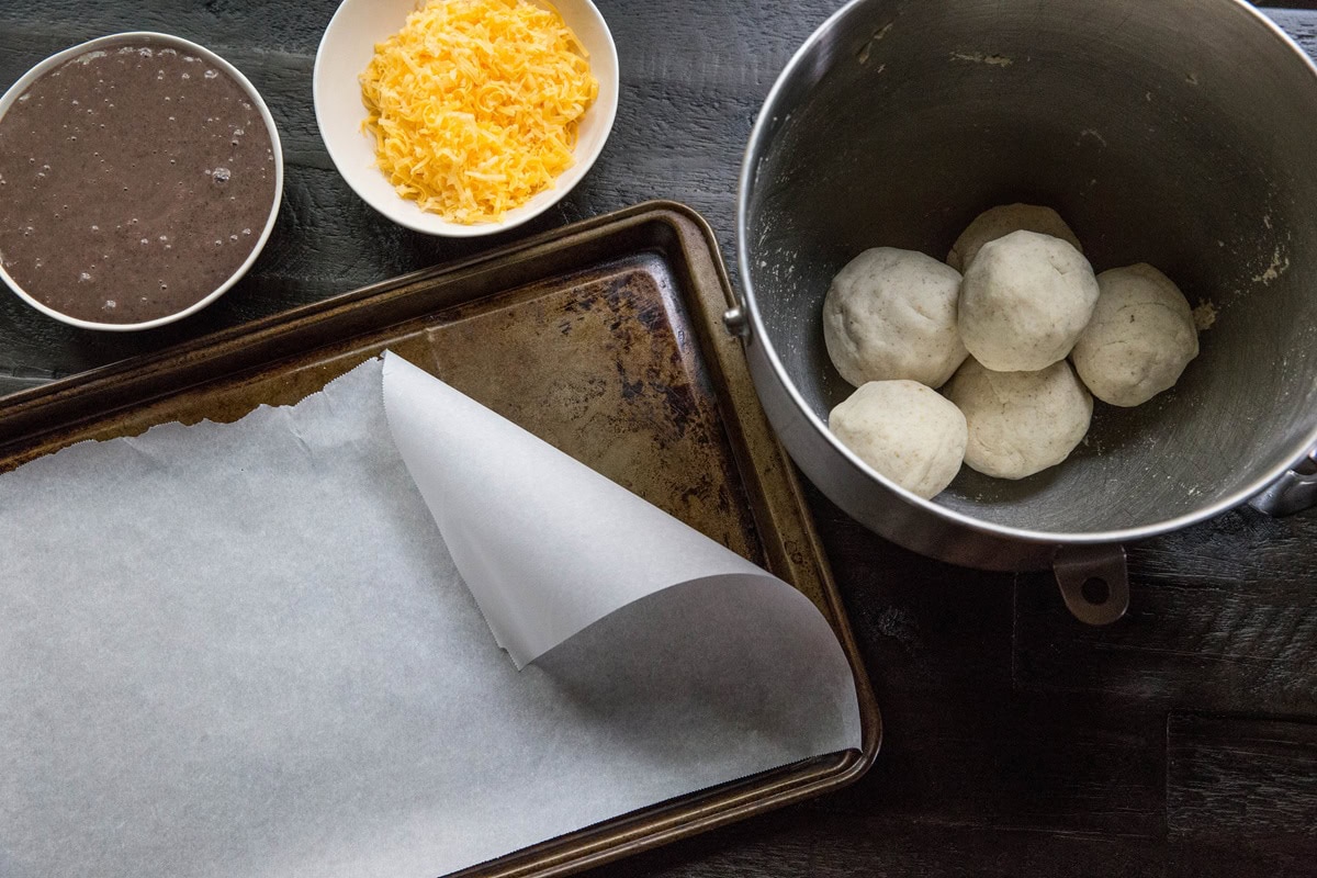 A baking tray lined with parchment paper, a bowl of shredded cheese, black bean paste, and six dough balls set the stage for making gluten-free cheese flatbread inspired by the classic pupusa recipe on a dark wooden surface.