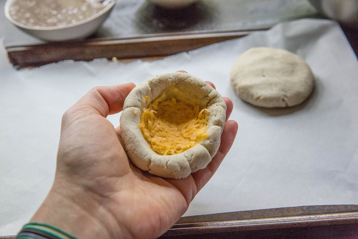 A hand holds an uncooked dough cup filled with shredded cheese, ready to become delicious cheese pupusas, with another dough round on parchment paper in the background.