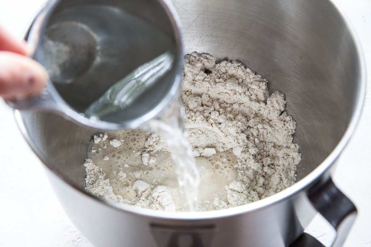 A metal mixing bowl containing flour, sugar, and yeast—key ingredients for a cheese pupusas or Gluten-Free Cheese Flatbread recipe—viewed from above on a light surface with a hand pouring water into the bowl of dry ingredients.
