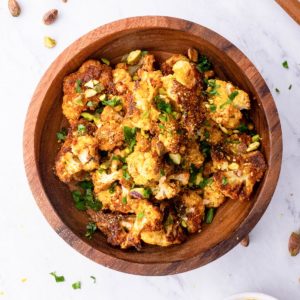 Wooden bowl full of vegan roasted cauliflower with nutritional yeast to the side and fresh parsley sprinkled around.