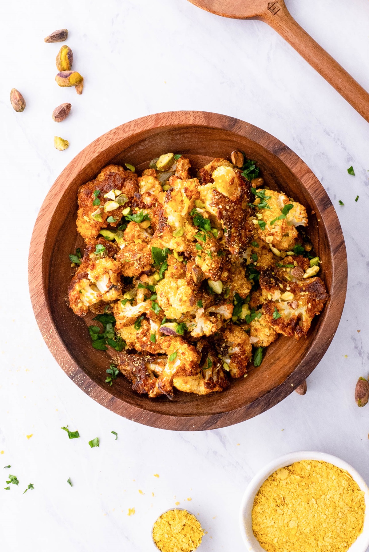 Wooden bowl full of vegan roasted cauliflower with nutritional yeast to the side and fresh parsley sprinkled around.