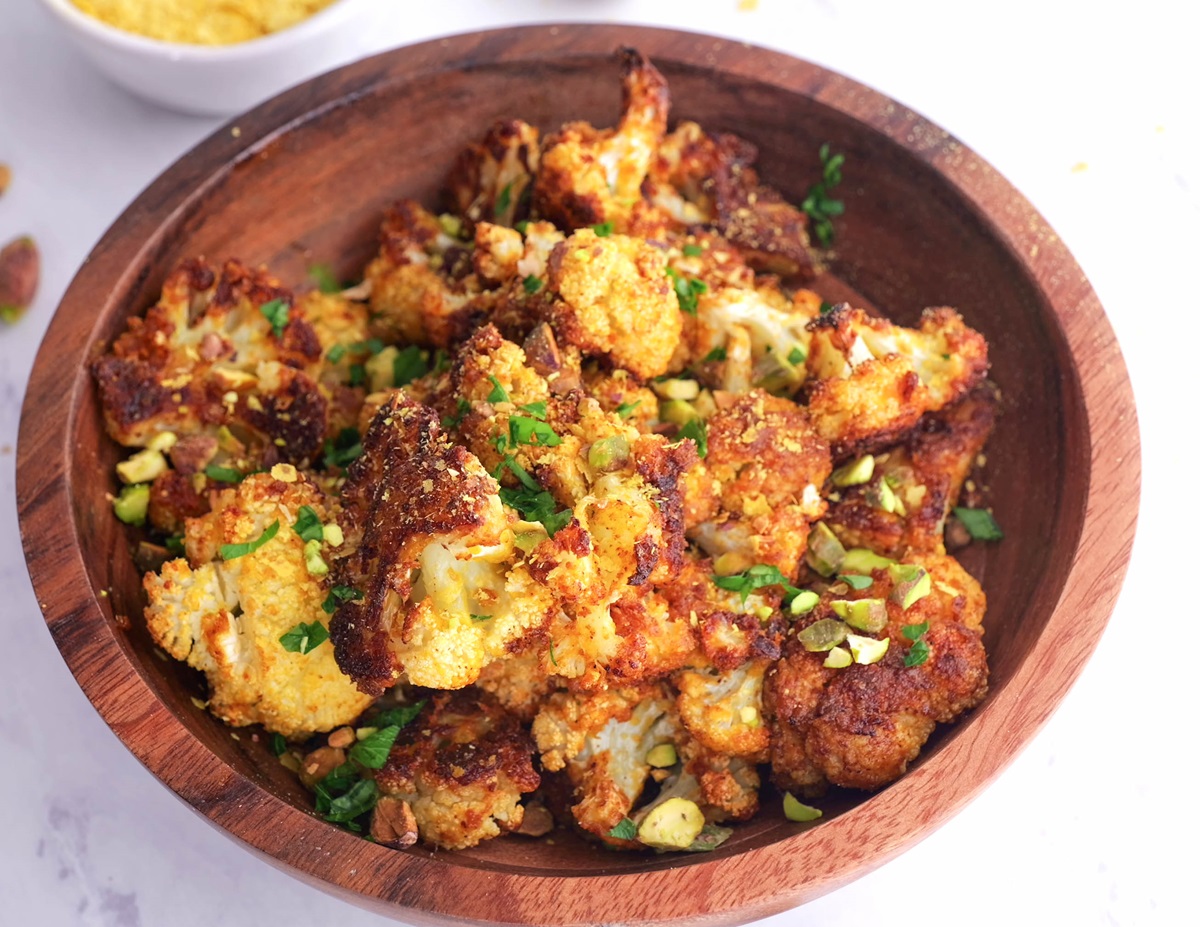 Wooden bowl with roasted cauliflower inside, fresh out of the oven. Pistachios, a bowl of nutritional yeast, and wooden spoons to the side.