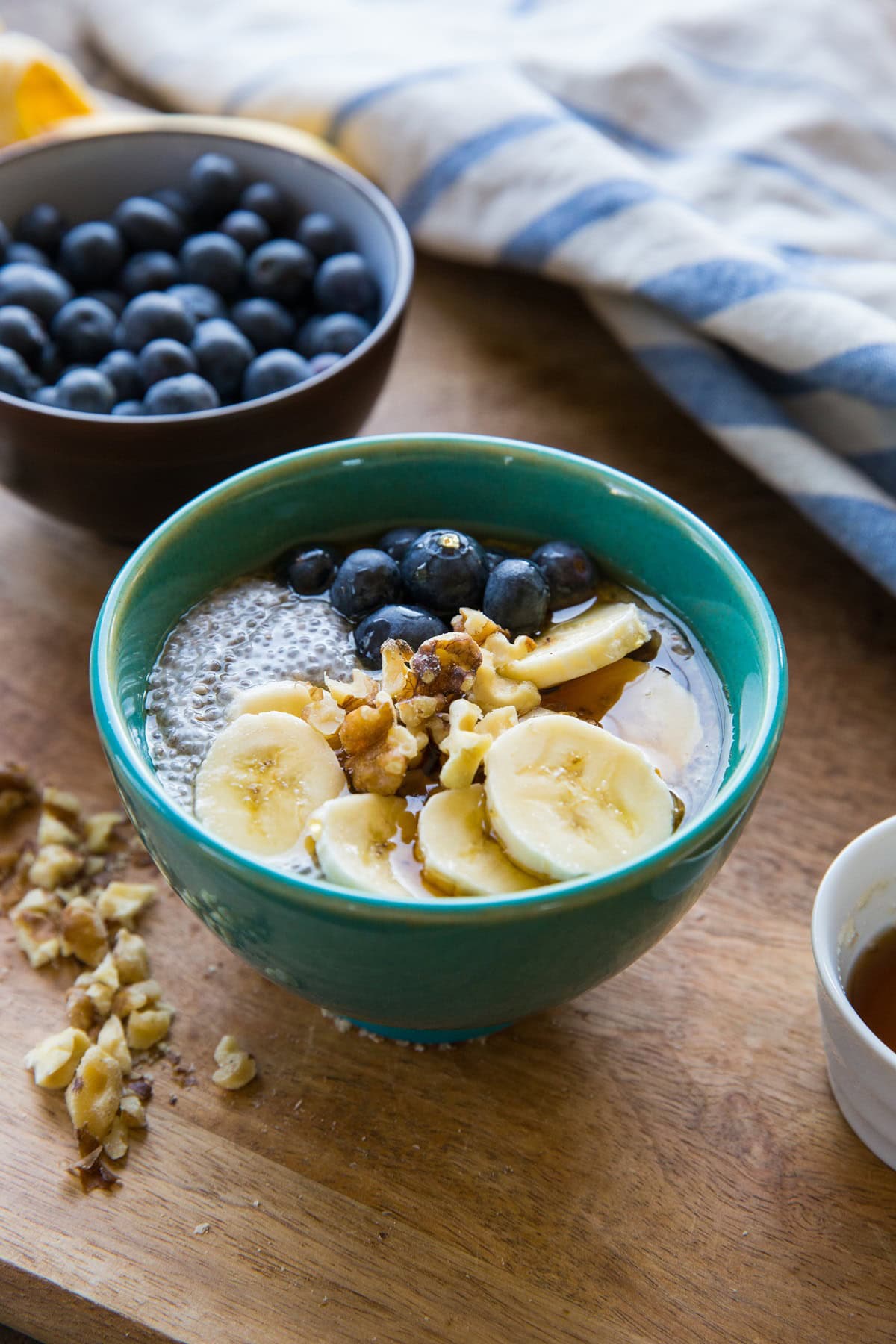 small teal bowl of chia pudding topped with fresh fruit, nuts, and a drizzle of maple syrup. Fresh blueberries and a napkin in the background.