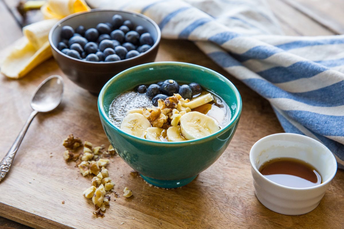 Teal bowl of chia seed pudding topped with sliced bananas, blueberries, walnuts and pure maple syrup. Small bowl of fresh blueberries in the background and a napkin