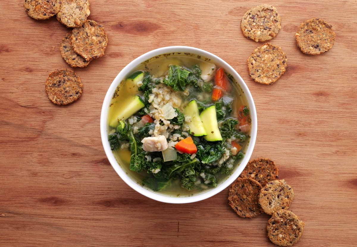 Bowl of chicken soup with rice and kale on a wooden background with crackers all around.