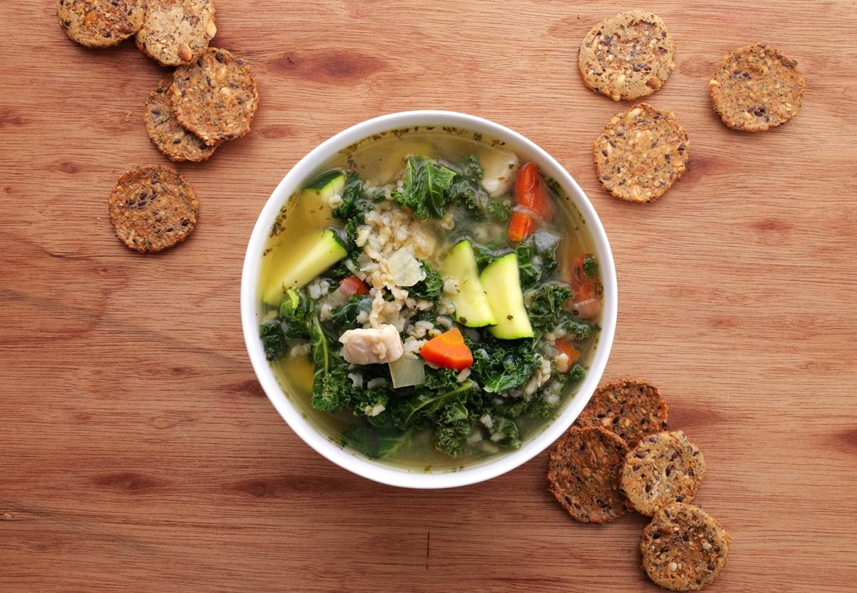 Bowl of chicken soup with rice and kale on a wooden background with crackers all around.