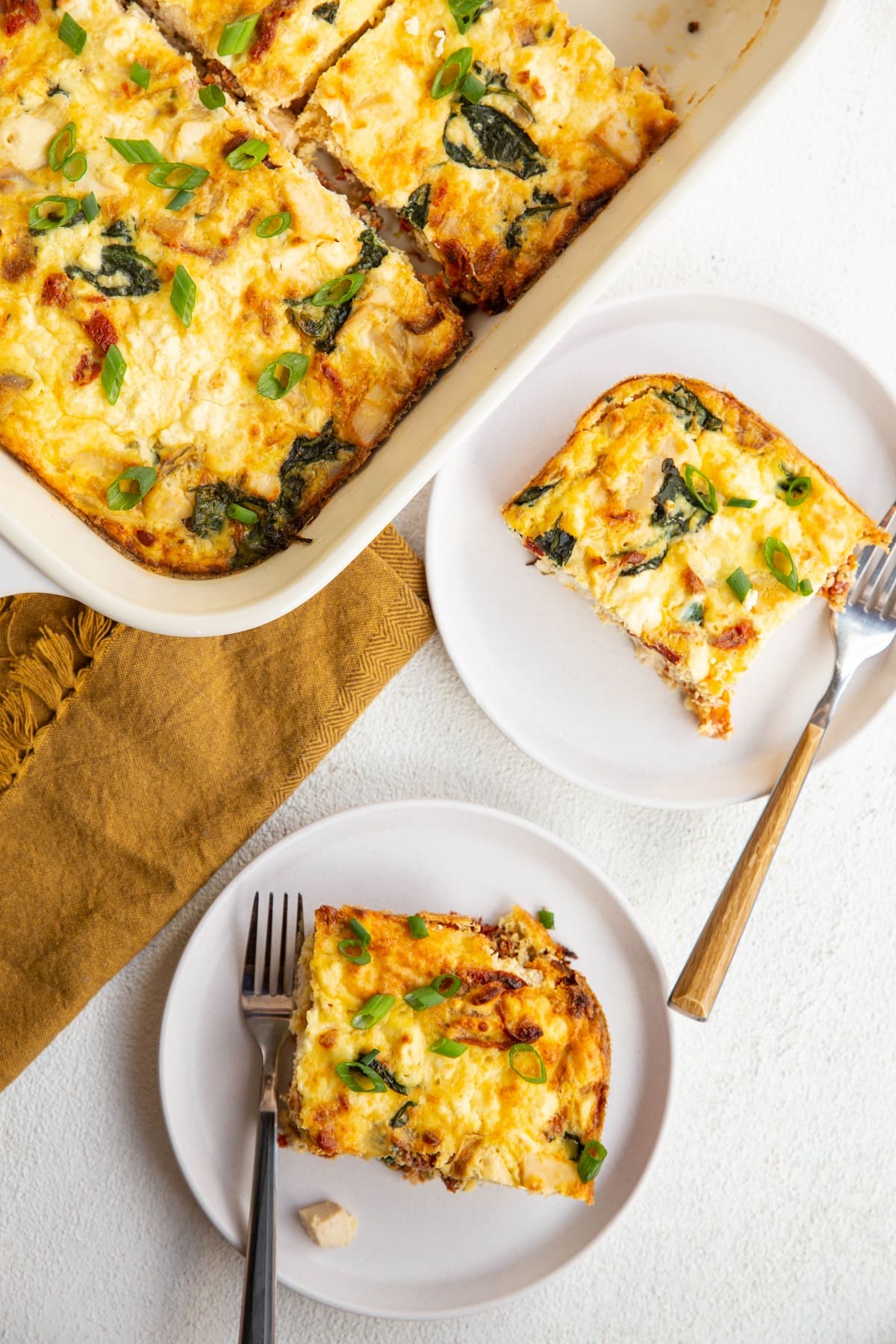 Two slices of egg casserole on white plates with the casserole dish of casserole to the side.