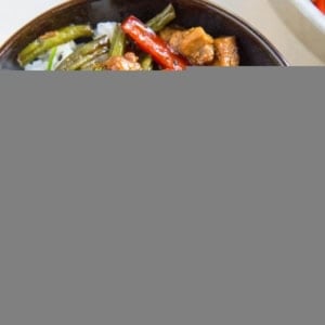 A bowl of white rice topped with Teriyaki Chicken, stir-fried green beans, and red bell peppers, garnished with chopped green onions and black sesame seeds. Chopsticks rest nearby on a light table.
