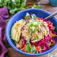 Blue bowl with burrito bowl ingredients inside including steamed rice, shredded chicken, avocado and purple cabbage slaw.
