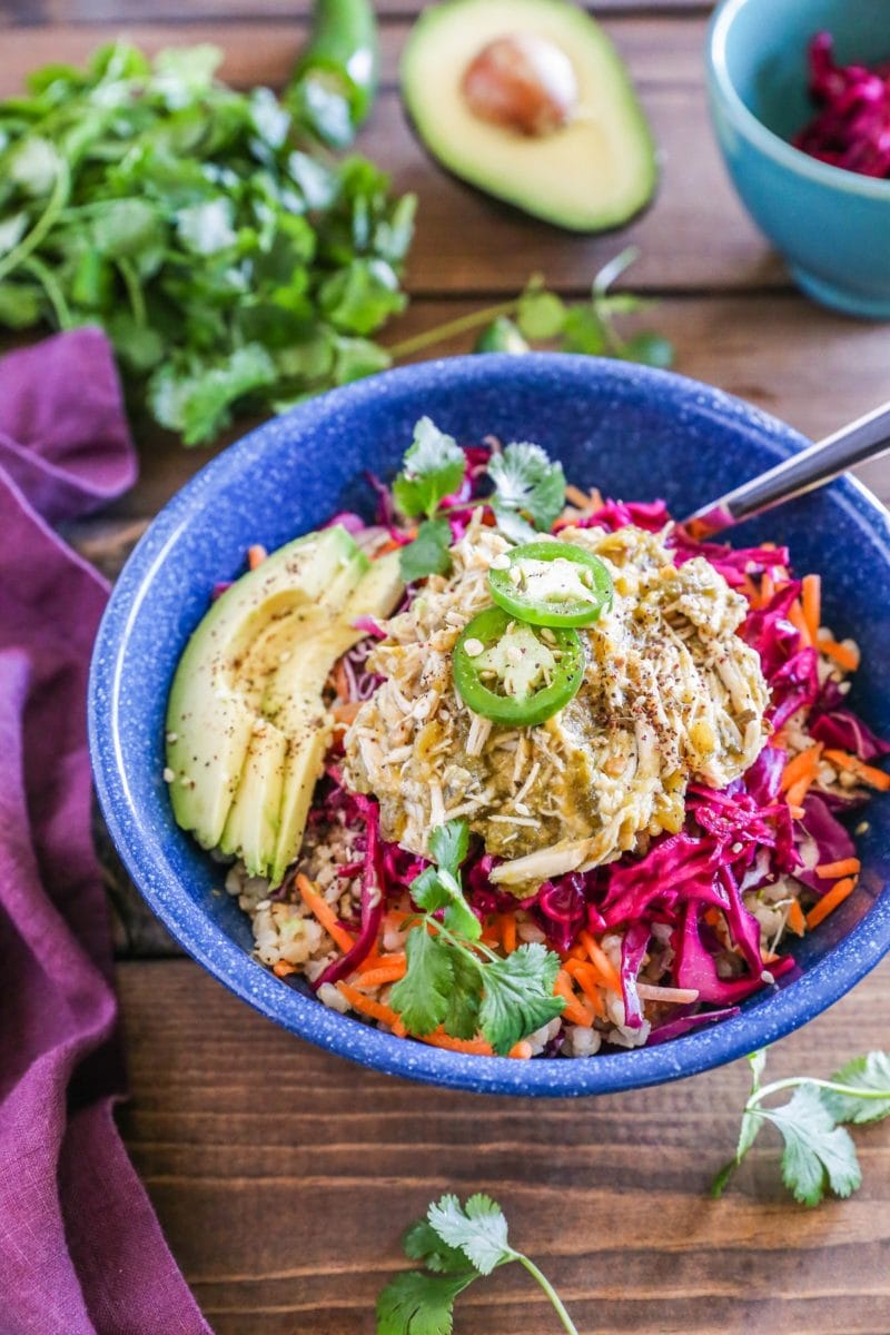 Blue bowl with burrito bowl ingredients inside including steamed rice, shredded chicken, avocado and purple cabbage slaw.