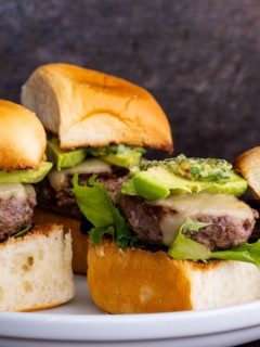 Beef sliders on a white plate, ready to serve. Topped with avocado slices and chimichurri sauce