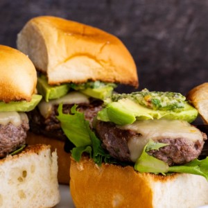 Beef sliders on a white plate, ready to serve. Topped with avocado slices and chimichurri sauce