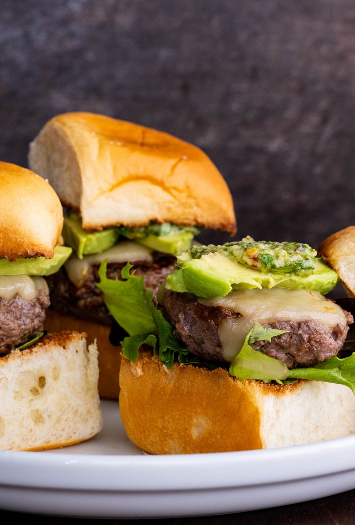 Beef sliders on a white plate, ready to serve. Topped with avocado slices and chimichurri sauce