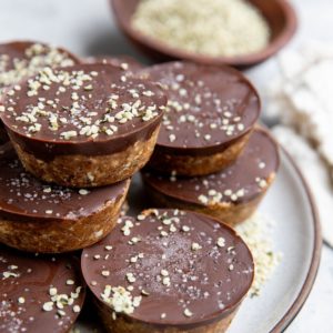Stack of chocolate covered protein energy cups on a plate, sprinkled with hemp seeds. A bowl of hemp seeds in the background.