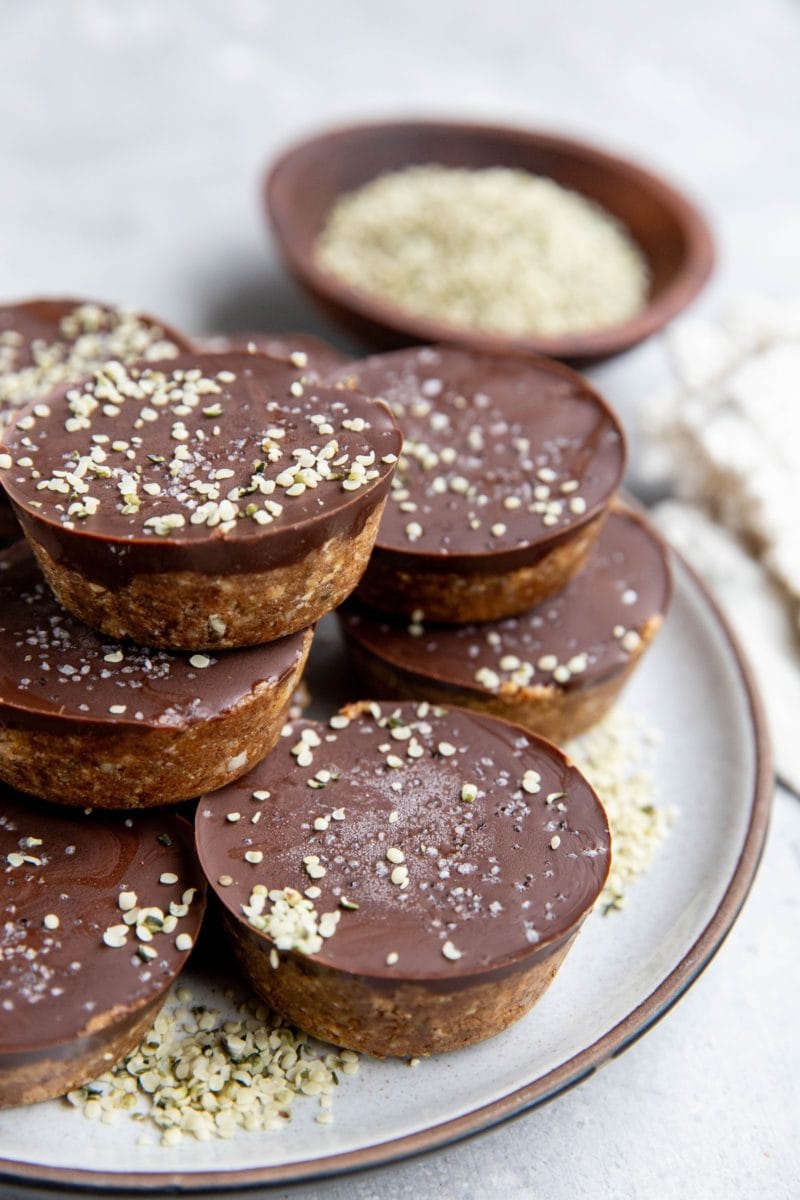 Stack of chocolate covered protein energy cups on a plate, sprinkled with hemp seeds. A bowl of hemp seeds in the background.