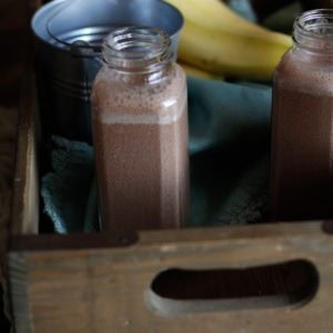 Two Chocolate Hemp Seed Smoothies in jars inside of a wooden crate with a light linen and fresh bananas. Ready to drink.