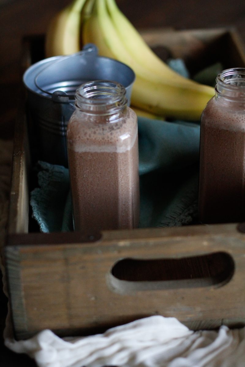 Two Chocolate Hemp Seed Smoothies in jars inside of a wooden crate with a light linen and fresh bananas. Ready to drink.