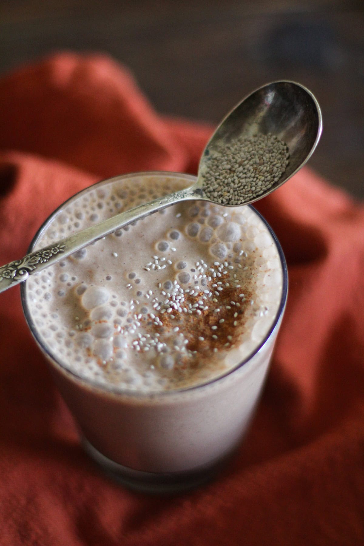 Glass full of chocolate peanut butter smoothie with chia seeds and cinnamon sprinkled on top with a spoon of chia seeds resting on top of the glass.
