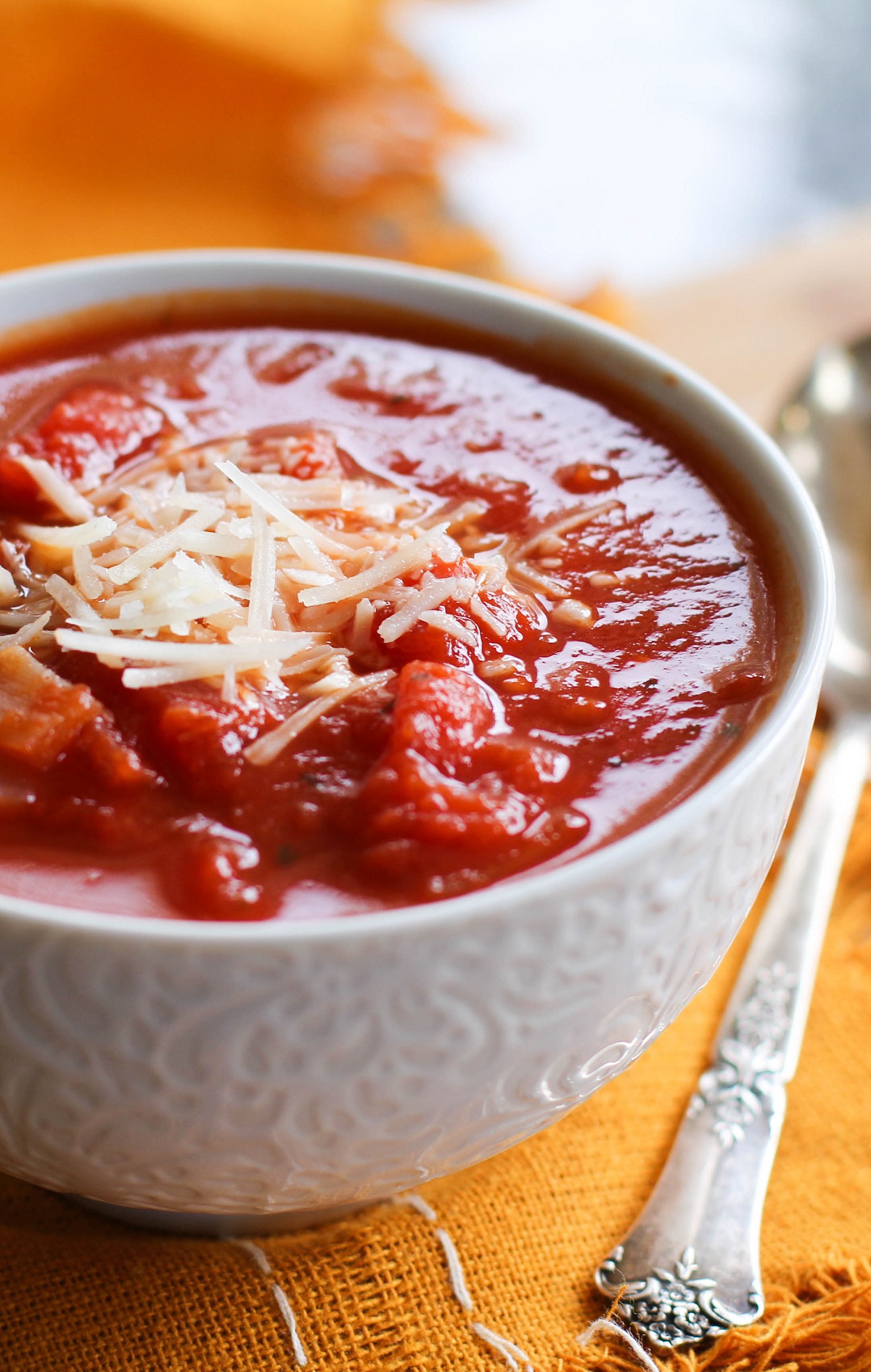 White bowl of chunky tomato basil soup with an orange napkin and an old fashioned spoon. Cheese sprinkled on top of the soup