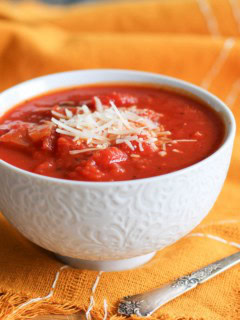 Small white bowl of chunky tomato soup with parmesan cheese on top and an orange napkin and silver spoon.