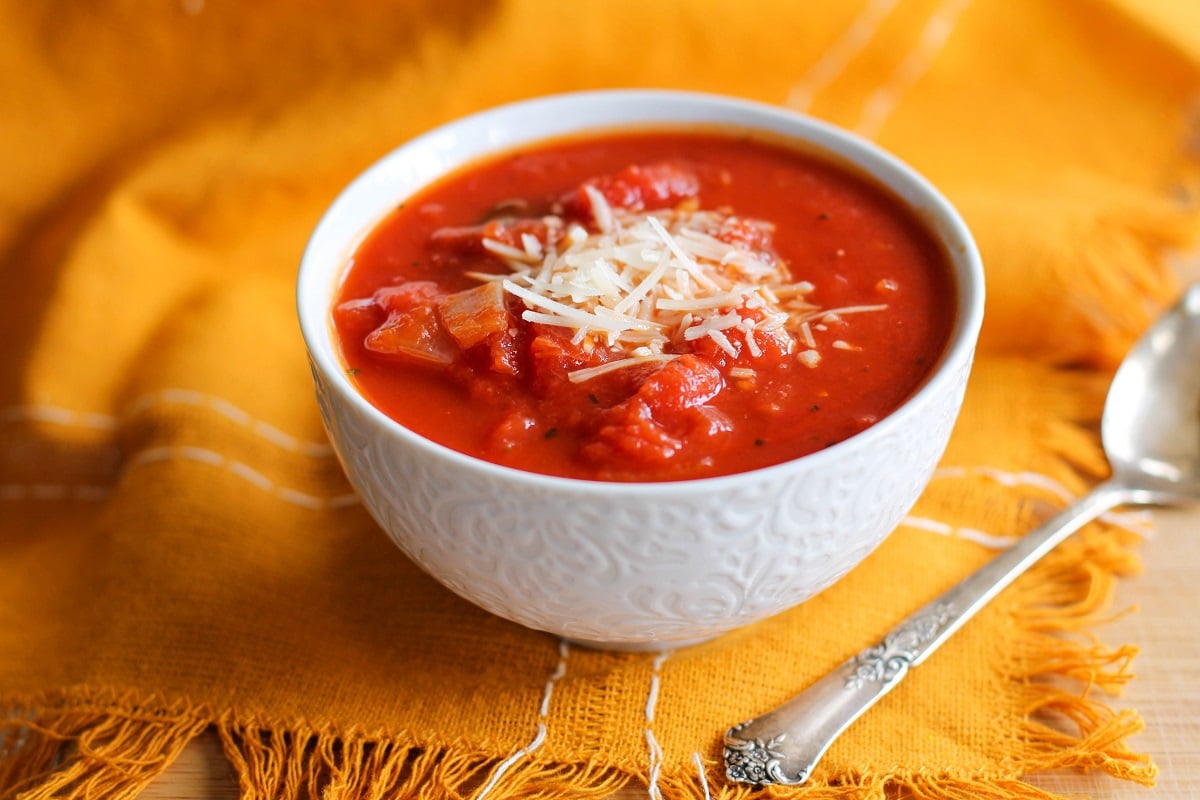 White bowl of tomato basil soup on a wooden cutting board with a napkin and a spoon.