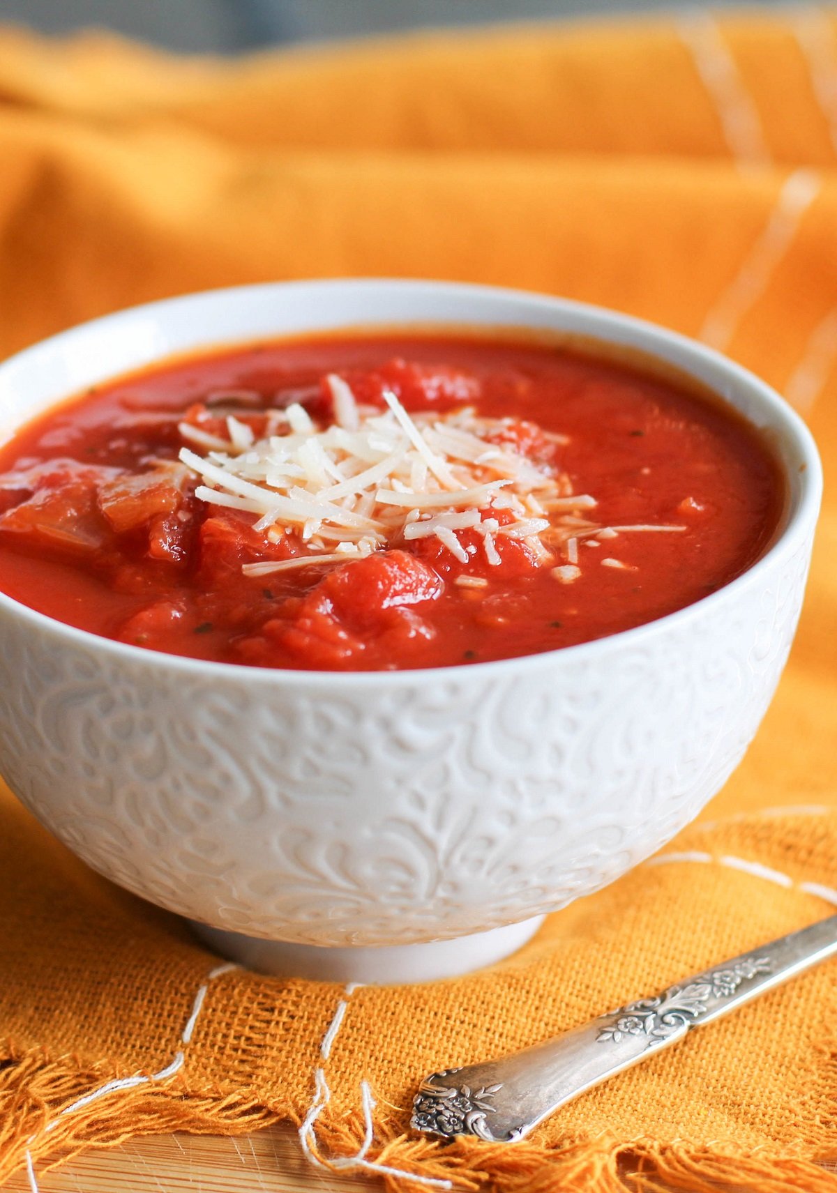 Small white bowl of chunky tomato soup with parmesan cheese on top and an orange napkin and silver spoon.