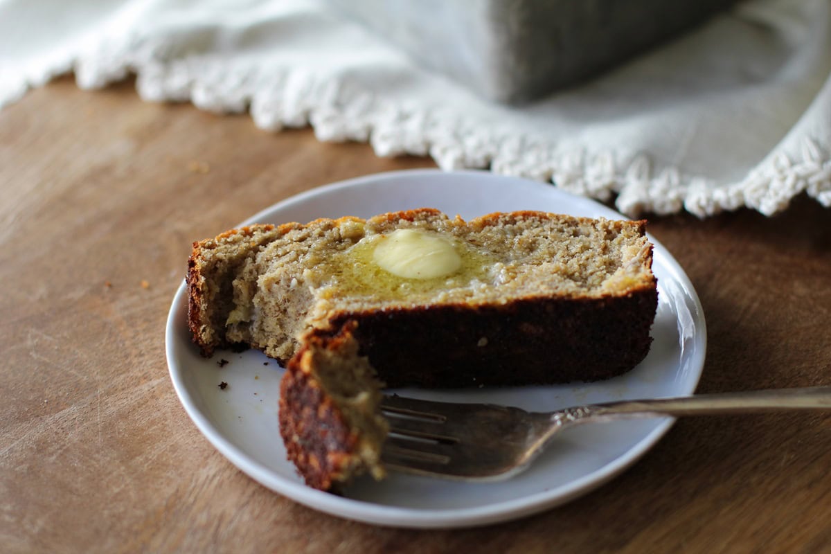 Thick slice of banana bread on a small plate with melted butter on top and a fork taking a bite out. A cream colored napkin in the background.