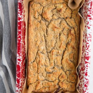A rectangular loaf of golden-brown coconut flour banana bread with a cracked top sits in a red and white speckled baking dish lined with brown parchment paper.