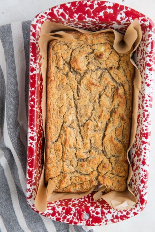 A rectangular loaf of golden-brown coconut flour banana bread with a cracked top sits in a red and white speckled baking dish lined with brown parchment paper.