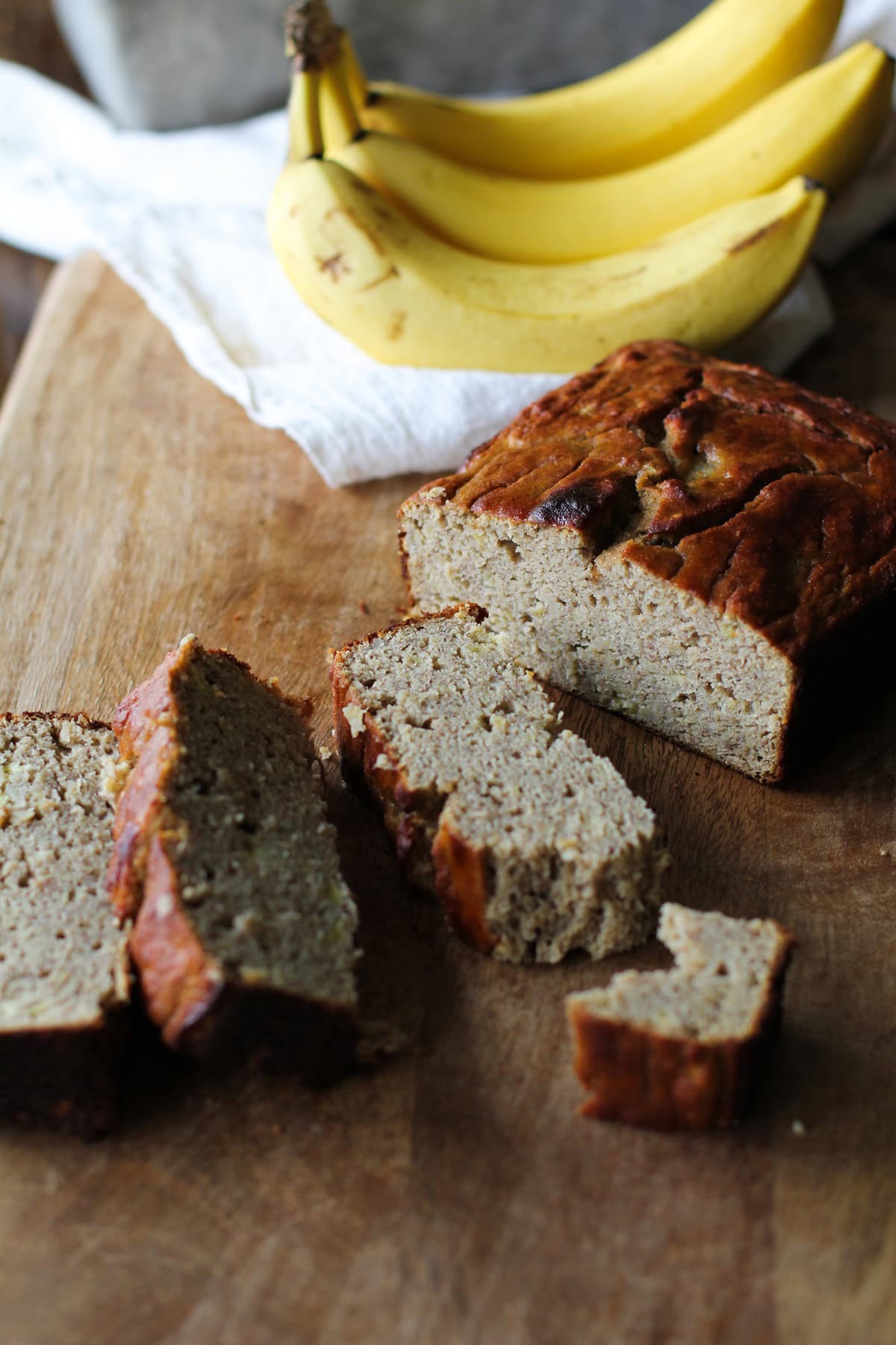 Loaf of grain-free banana bread on a cutting board cut into slices with a bite torn off of one of the pieces. Fresh bananas and a napkin in the background.