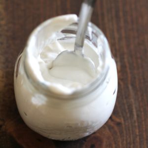 A glass jar filled with creamy white yogurt sits on a wooden surface, topped with a swirl of coconut whipped cream, with a metal spoon inside. A single red cherry lies nearby on the table.