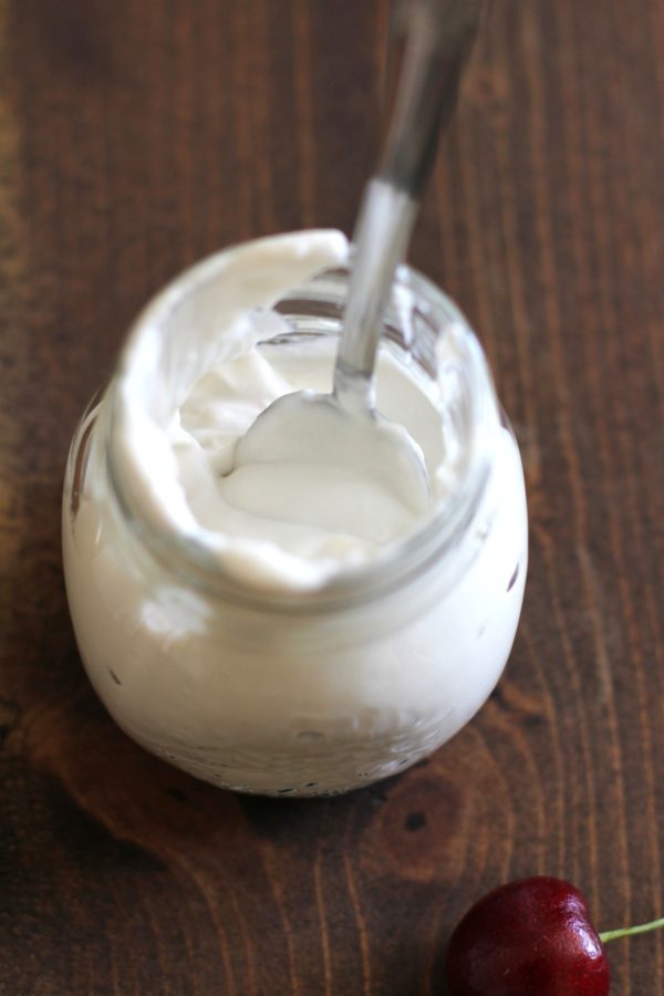 A glass jar filled with creamy white yogurt sits on a wooden surface, topped with a swirl of coconut whipped cream, with a metal spoon inside. A single red cherry lies nearby on the table.