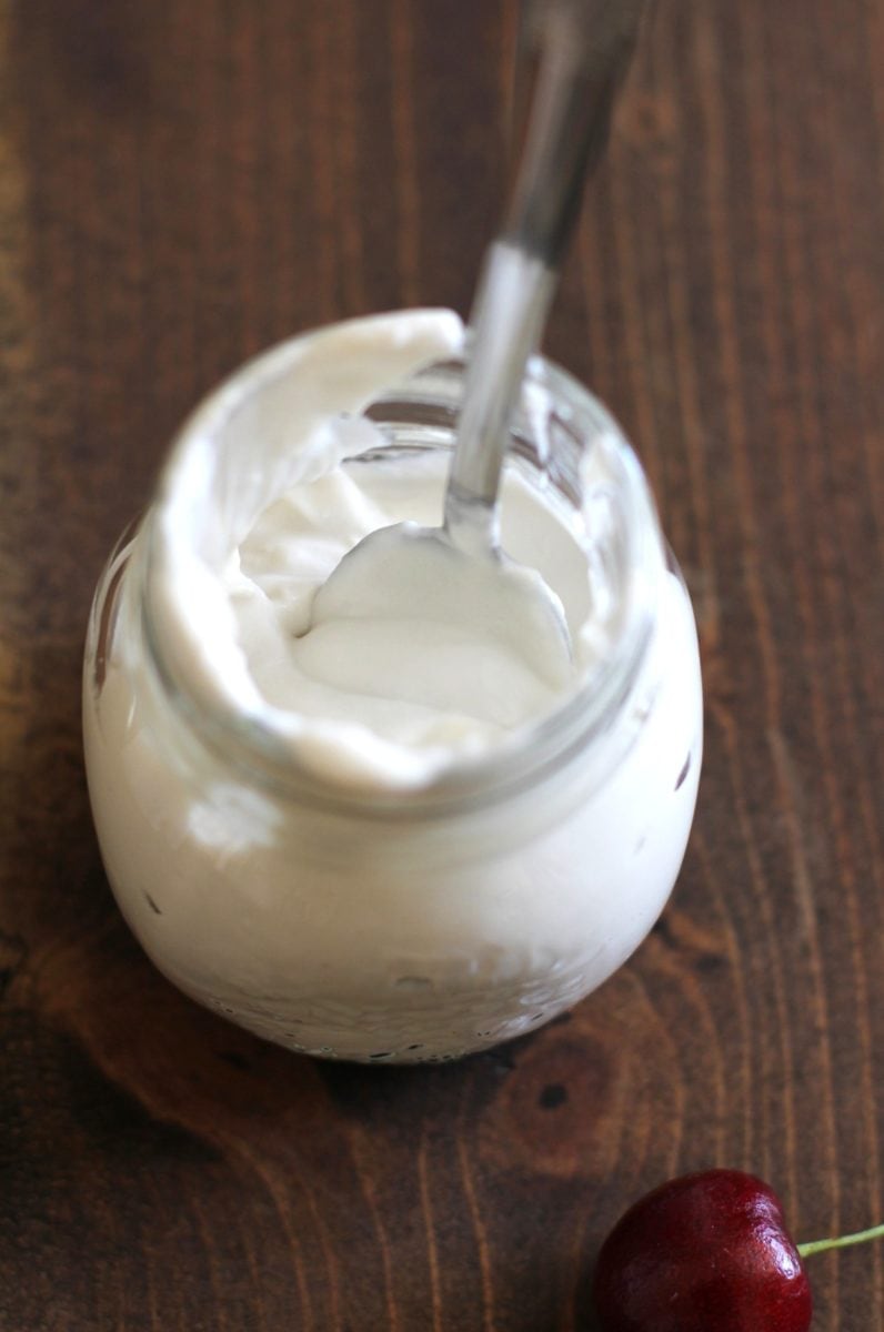 A glass jar filled with creamy white yogurt sits on a wooden surface, topped with a swirl of coconut whipped cream, with a metal spoon inside. A single red cherry lies nearby on the table.
