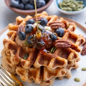 Three waffles stacked on a plate topped with almond butter, blueberries, and pecans with honey being poured on top. A small bowl of blueberries and a small bowl of pumpkin seeds in the background. There's a golden fork next to the waffles and a golden napkin to the side.