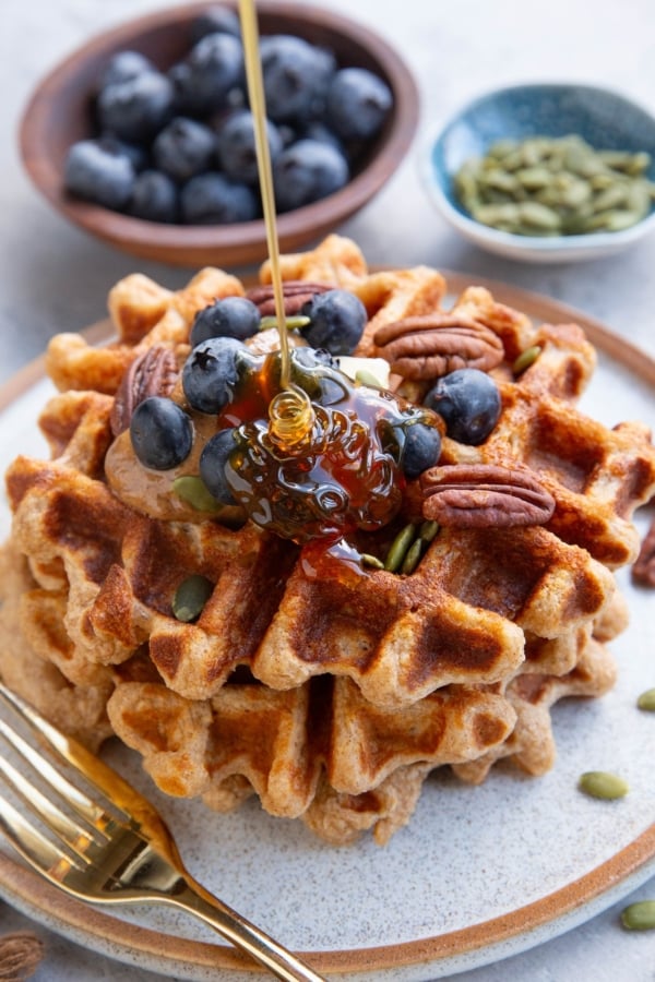 Three waffles stacked on a plate topped with almond butter, blueberries, and pecans with honey being poured on top. A small bowl of blueberries and a small bowl of pumpkin seeds in the background. There's a golden fork next to the waffles and a golden napkin to the side.