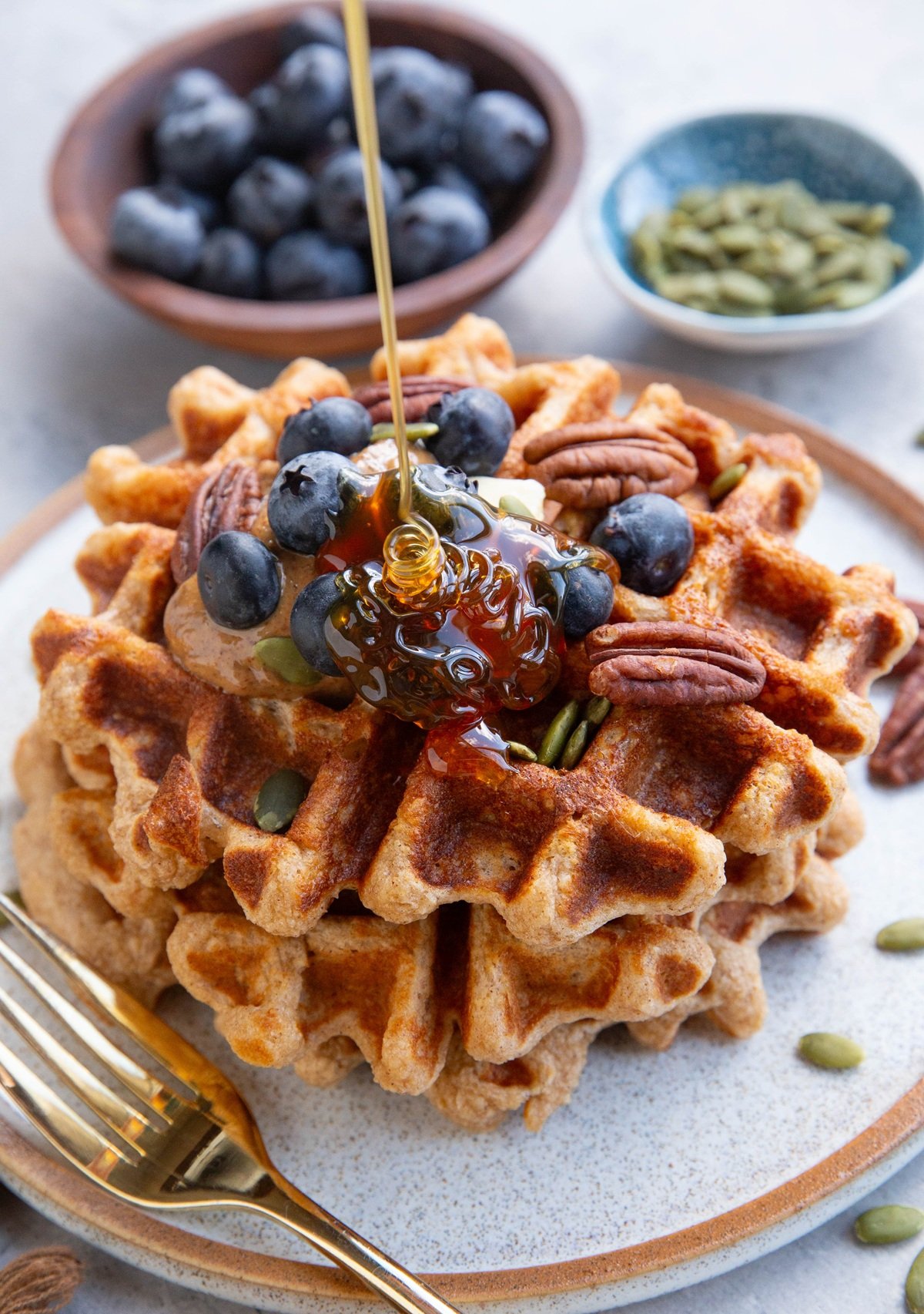 Three waffles stacked on a plate topped with almond butter, blueberries, and pecans with honey being poured on top. A small bowl of blueberries and a small bowl of pumpkin seeds in the background. There's a golden fork next to the waffles and a golden napkin to the side.