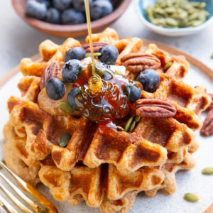Three waffles stacked on a plate topped with almond butter, blueberries, and pecans with honey being poured on top. A small bowl of blueberries and a small bowl of pumpkin seeds in the background. There's a golden fork next to the waffles and a golden napkin to the side.