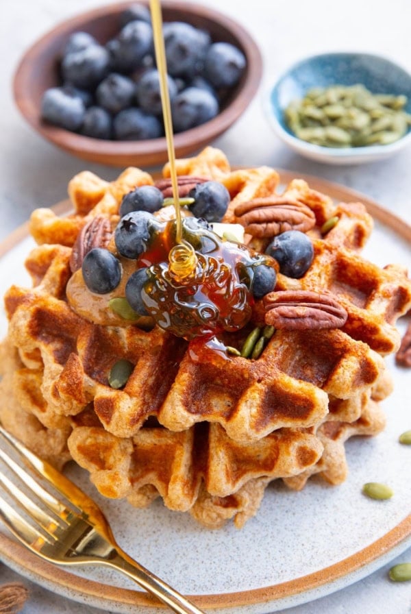 Three waffles stacked on a plate topped with almond butter, blueberries, and pecans with honey being poured on top. A small bowl of blueberries and a small bowl of pumpkin seeds in the background. There's a golden fork next to the waffles and a golden napkin to the side.