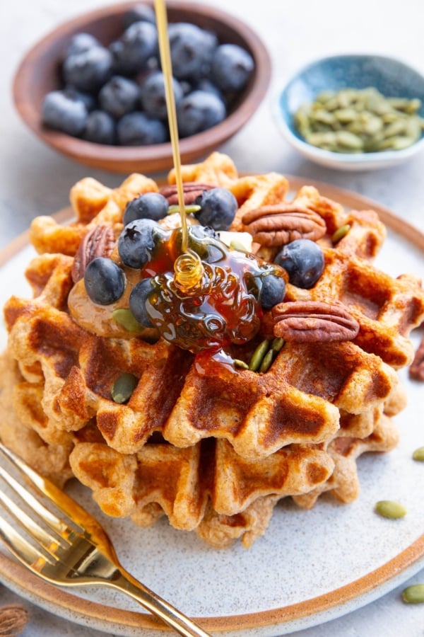 Three waffles stacked on a plate topped with almond butter, blueberries, and pecans with honey being poured on top. A small bowl of blueberries and a small bowl of pumpkin seeds in the background. There's a golden fork next to the waffles and a golden napkin to the side.